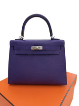 A structured purple top-handle handbag with silver hardware, displayed on an orange box against a white background.