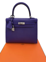 A structured purple top-handle handbag with silver hardware, displayed on an orange box against a white background.