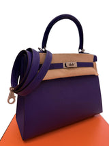 A structured purple leather handbag with a tan leather top band, a matching purple shoulder strap draped over the side, and silver-tone hardware, photographed on a white background with an orange surface visible beneath the bag.