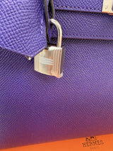 Close-up of a purple textured leather handbag with a silver padlock and a small Hermes Paris logo printed on an orange surface at the bottom right.