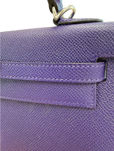 Close-up of a purple textured leather handbag showing the strap tab, stitching, and part of a dark handle with a metal ring on a white background.