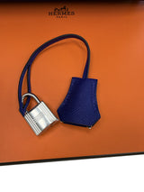 A blue textured leather Hermes key fob with a silver padlock, displayed on an orange Hermes box background.